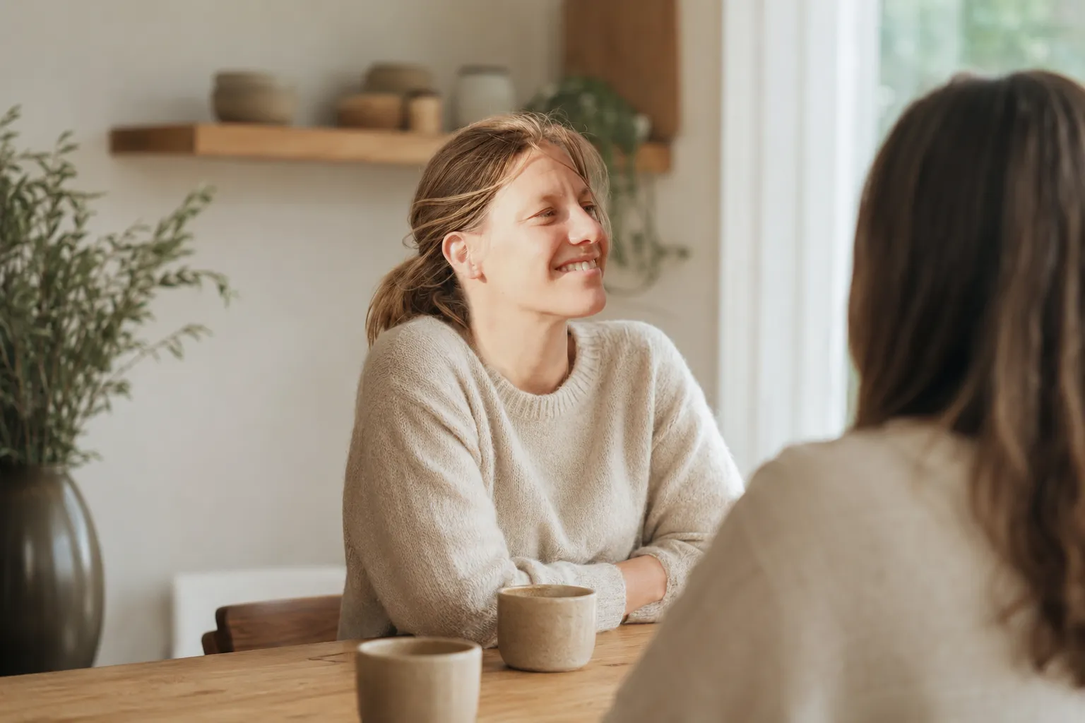 Zwei Hände halten eine Keramiktasse an einem hölzernen Tisch, gegenüber sitzt eine zweite Person mit ihrer Tasse.